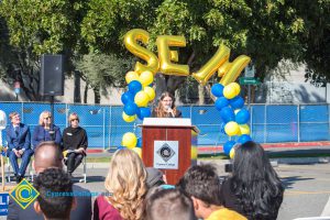 Young lady with long brown hair and glasses speaking to audience at SEM Groundbreaking ceremony.