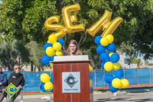 Young woman with glasses at podium with blue and yellow balloon arch and SEM balloons.