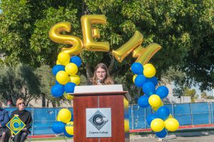 Young woman with glasses at podium with blue and yellow balloon arch and SEM balloons.