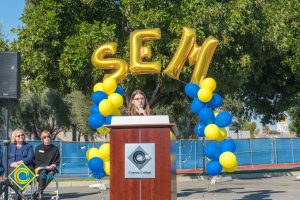 Young woman with glasses at podium with blue and yellow balloon arch and SEM balloons.