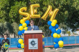 Man in a suit at podium with blue and yellow balloon arch and SEM balloons.