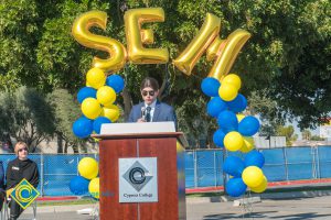 Man in a suit at podium with blue and yellow balloon arch and SEM balloons.