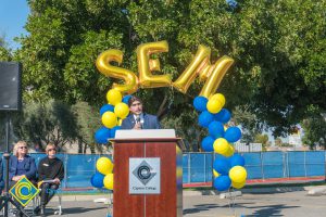 Man in a suit at podium with blue and yellow balloon arch and SEM balloons.