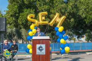 Man in a suit at podium with blue and yellow balloon arch and SEM balloons.