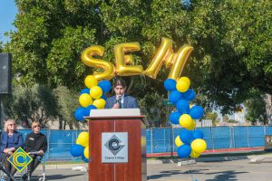Man in a suit at podium with blue and yellow balloon arch and SEM balloons.