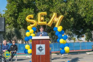 Man in a suit at podium with blue and yellow balloon arch and SEM balloons.