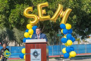 Man in a suit at podium with blue and yellow balloon arch and SEM balloons.