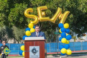 Man in a suit at podium with blue and yellow balloon arch and SEM balloons.