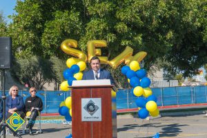 Man in a suit at podium with blue and yellow balloon arch and SEM balloons.