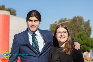 Young man with blue suit and tie with his arm around a young woman with glasses and long brown hair.