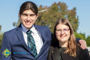 Young man with blue suit and tie with his arm around a young woman with glasses and long brown hair.