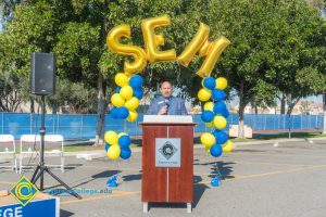 Marc Posner at podium with blue and yellow balloon arch and SEM balloons.