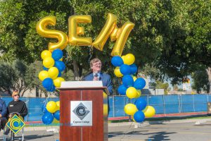 Richard Fee at podium with blue and yellow balloon arch and SEM balloons.