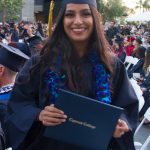 A young lady with long dark hair with a blue lei smiling in her cap and gown.