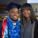 A male graduate in his cap and gown with a blue floral lei and a woman with dark brown hair, wearing a grey scarf around her neck.