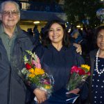 A smiling young lady in her cap and gown, holding a bouquet of flowers in each hand with a man and woman at her side,