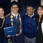 A graduate wearing his cap and gown and displaying his degree, surrounded by his family.
