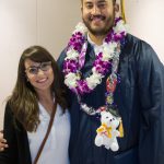 A graduate wearing full graduation regalia and floral lei with teddy bear with a smiling young woman with glasses.