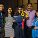 A graduate wearing her cap and gown, displaying her degree while holding a bouquet of sunflowers, surrounded by her family.