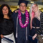 A graduate in cap and gown wearing a purple lei with two smiling women by his side.