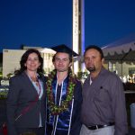 A graduate in cap and gown wearing a purple and green lei with a woman and man by his side.