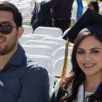 A young lady in her graduation cap sitting in a chair with a young man with a beard and moustache wearing sunglasses.