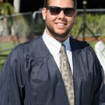 A smiling young man in sunglasses and beard wearing his graduation gown and tie.