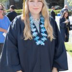 A young woman with blonde hair wearing her cap and gown and blue floral lei.
