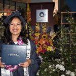 Smiling young lady in cap and gown, holding her degree in front of the floral decor.