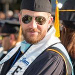 Young man with sunglasses and beard wearing cap and gown during commencement.