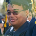 A young man wearing sunglasses, looking at the camera in his cap and gown.