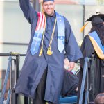 A young man in cap and gown holding up arm with his degree.