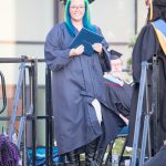 A young lady with blue and green hair and glasses in cap and gown during commencement.