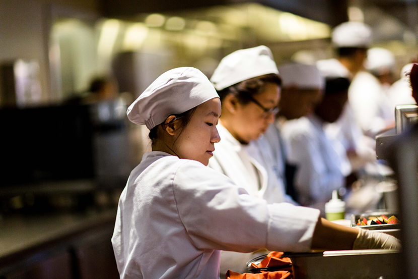 Two female culinary students in full uniform prepping food in the kitchen.