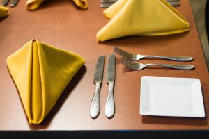 Table setting with gold, folded napkins, two knives, two forks and a white square plate.