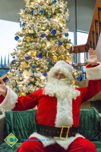 Santa Claus sitting with hands out and a large Christmas tree in the background with blue and gold ornaments.