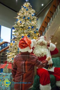 A very large Christmas tree with Santa Claus fixing his hat as someone stands at his side.