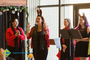 Woman wearing reindeer ears and a black scarf with Christmas lights speaking at the microphone while three other women look on.