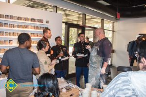 Three men in their Dress Blue uniform speak to a bearded man wearing a clay splattered apron while others look on.