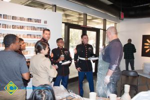 Three men in their Dress Blue uniform speak to a bearded man wearing a clay splattered apron while others look on.