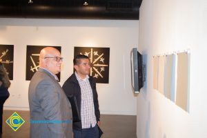 Juan Garcia and a man in a grey suit looking at various pieces of fabric displayed on the Art Gallery wall for the 100th Veteran's Day Anniversary.