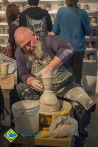 A bearded man with a clay spattered apron shapes a vase on a pottery wheel.