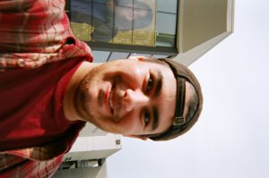 Sideway photo of a young man smiling with a backwards cap on his head.