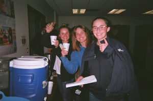 Three young ladies smiling next to drink cooler.