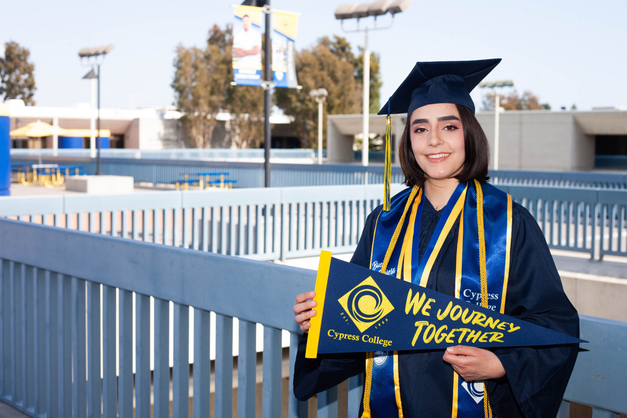 Portrait of Sabrina Tosti, the 2023 Presidential Scholar of Distinction for the CTE pathway, wearing regalia, holding a Cypress College "We Journey Together" pennant, with the 2nd floor piazza in the background.