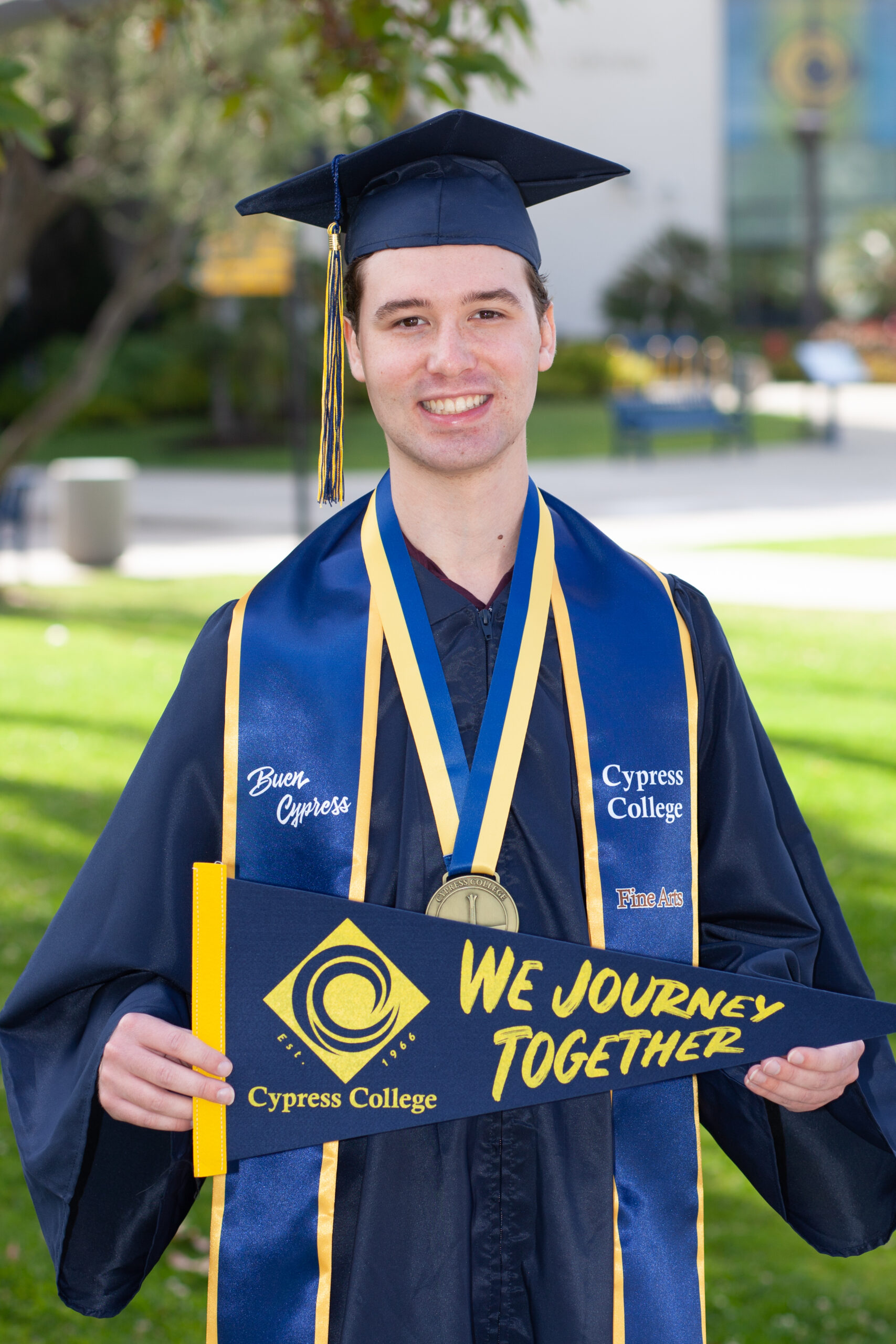 Portrait of Preston Harris, the 2023 Presidential Scholar of Distinction for the Fine Arts pathway, wearing regalia and holding a Cypress College "We Journey Together" pennant with Gateway Plaza, including the Student Center and Campanile, in the background.