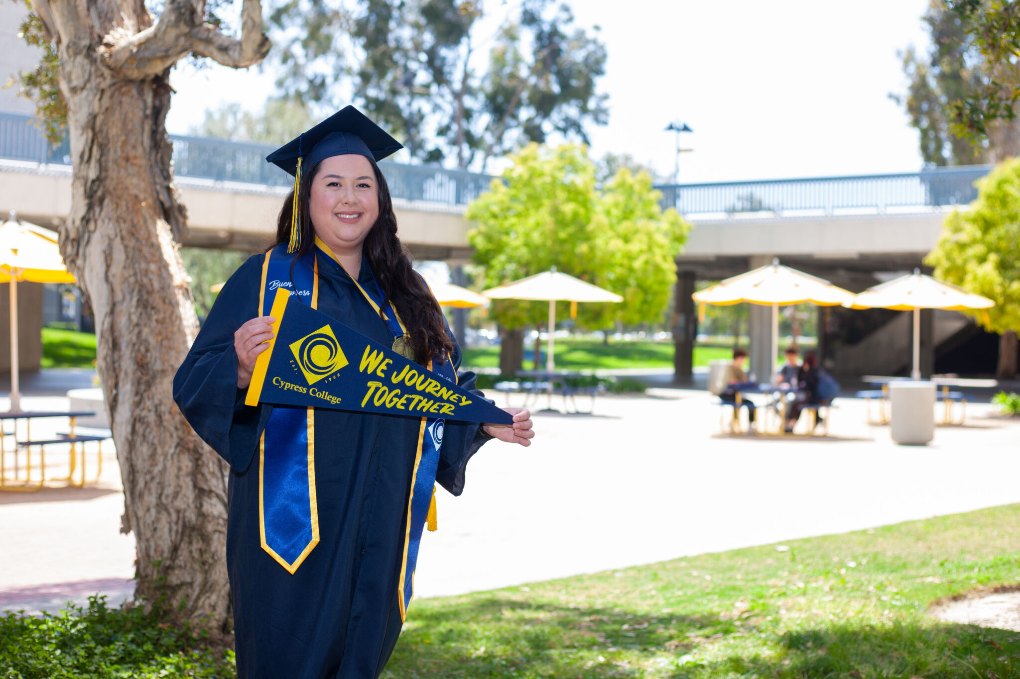 Portrait of Stephanie Wood, the 2023 Presidential Scholar of Distinction for the Health Science pathway, wearing regalia and holding a Cypress College "We Journey Together" pennant with the Fine Arts Plaza and piazza in the background.