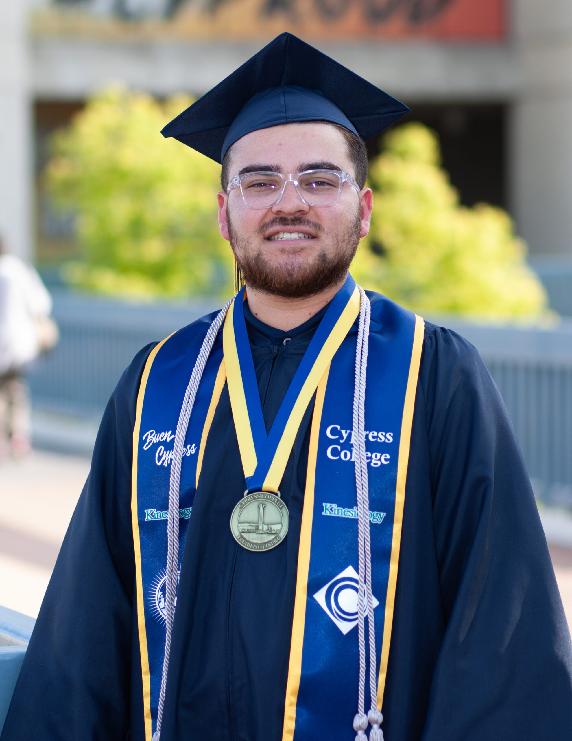 Portrait of Stephanie Wood, the 2023 Presidential Scholar of Distinction for the Health Science pathway, wearing regalia with the "#CYProud" window graphic in the background on the piazza near CCCPLX and Fine Arts.