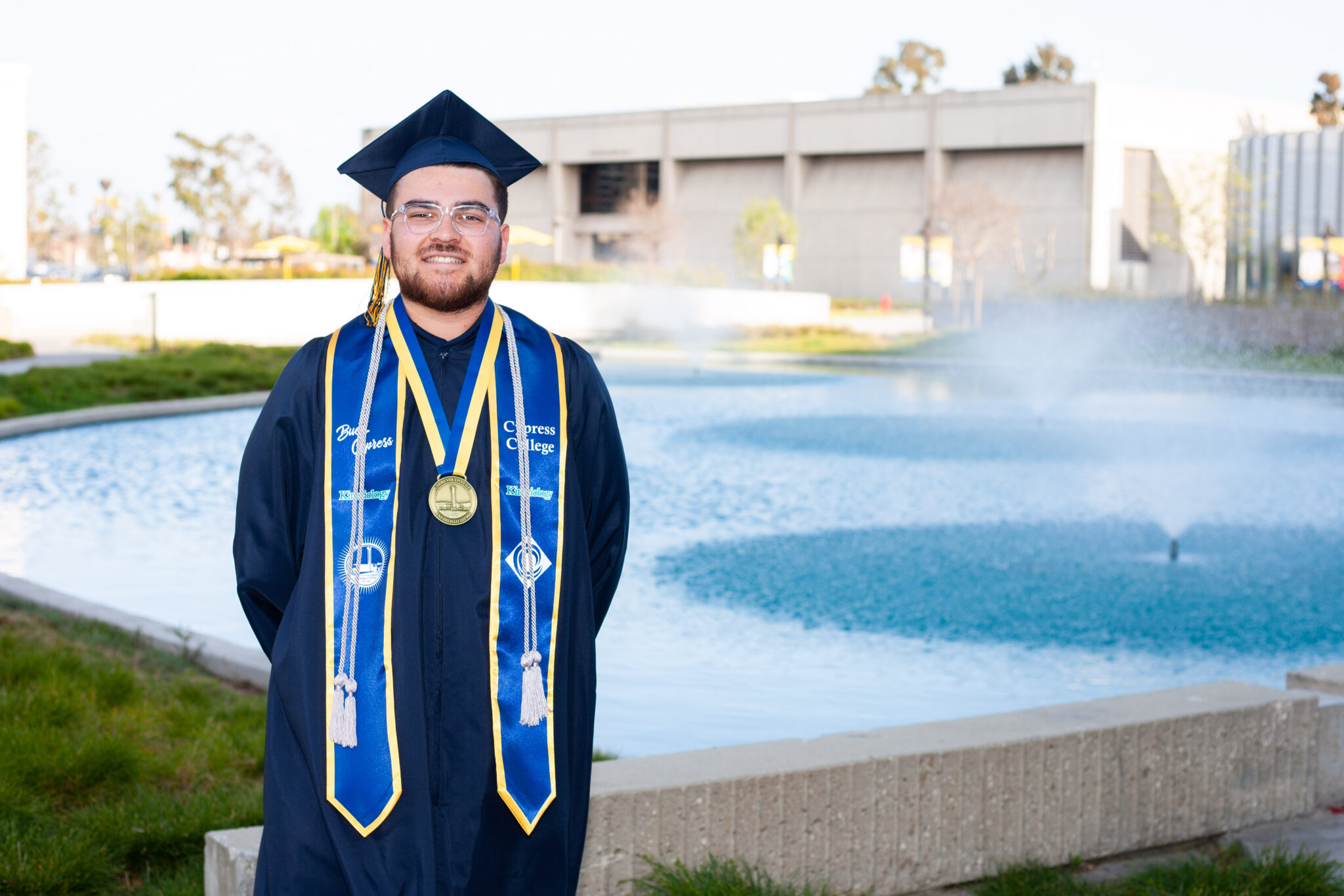 Portrait of Anthony Delgado, the 2023 Presidential Scholar of Distinction for the Kinesiology pathway, wearing regalia with the pond in the background.