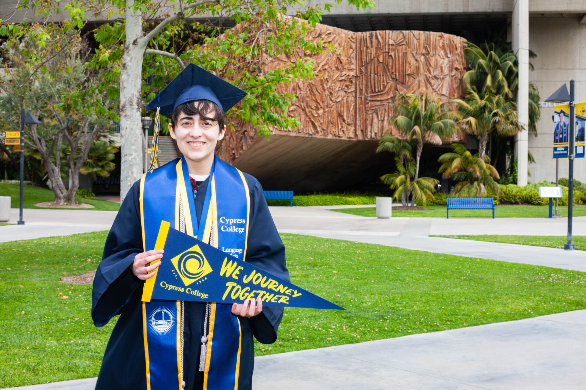 Portrait of Olly Tetrault, the 2023 Presidential Scholar of Distinction for the Language Arts pathway, wearing regalia and holding a Cypress College "We Journey Together" pennant with CCCPLX and the Sergio O'Cadiz sculpture in the background.